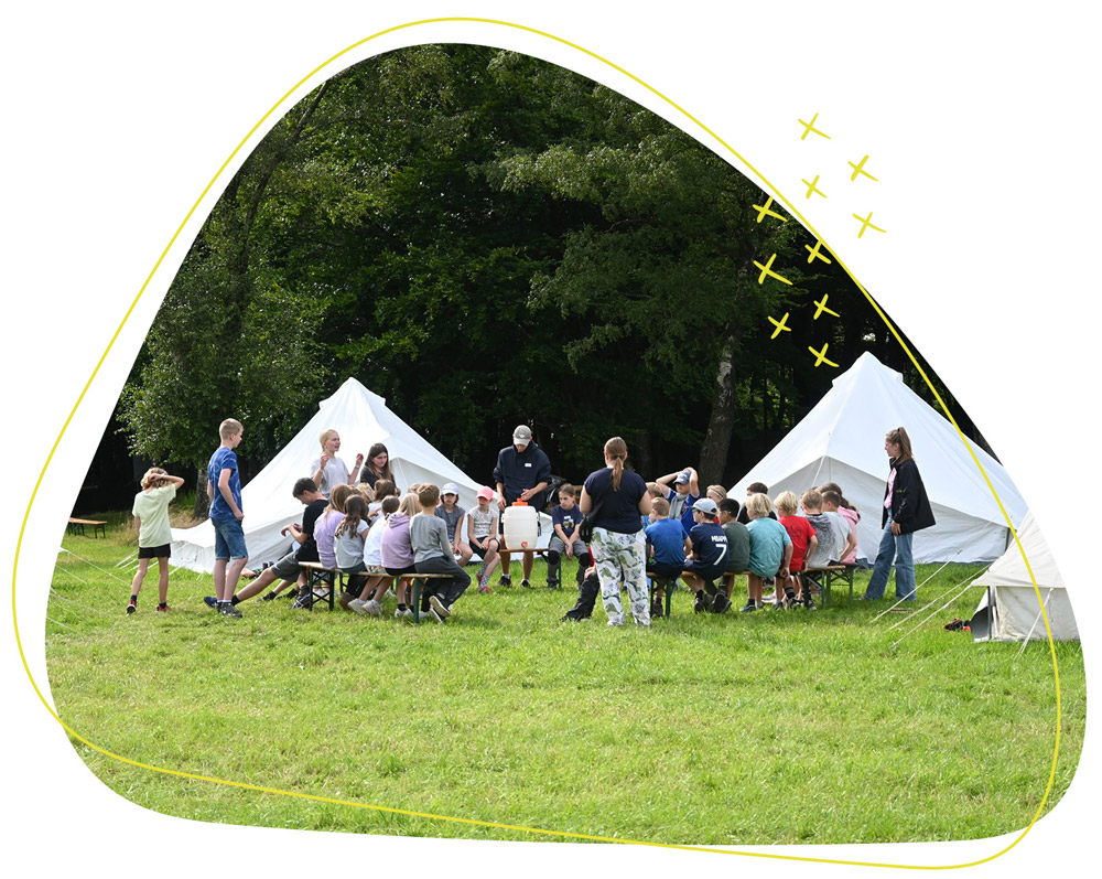Kinder sitzen in einem runden Kreis auf Bierbänken. Im Hintergrund sind Zelte