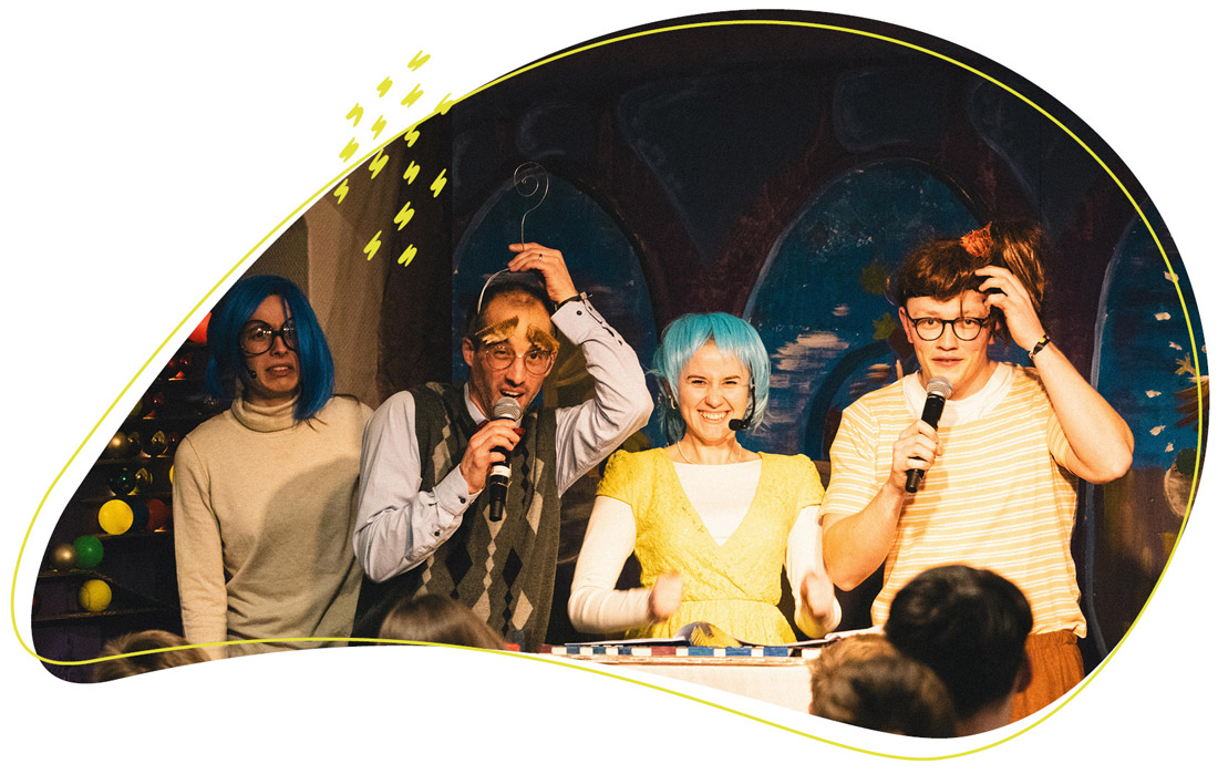 Auf dem Bild stehen vier bunt gekleidete Personen mit auffälligen Perücken und Mikrofonen auf einer kleinen Bühne und unterhalten ein Publikum in fröhlicher Atmosphäre.